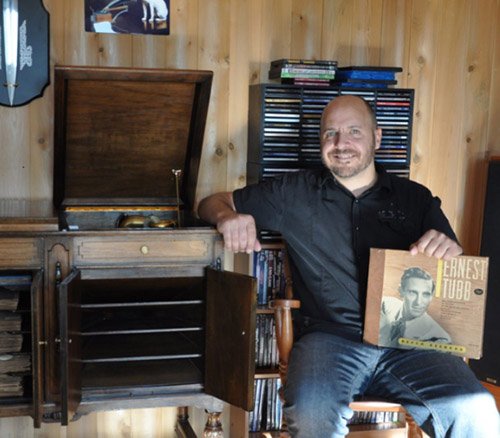 Homme accoudé à un meuble audio en bois avec gramophone, tenant une pochette de disque d'Ernest Tubb