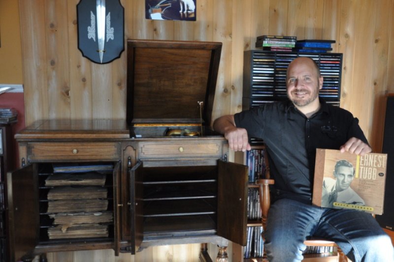 Homme accoudé à un meuble audio en bois avec gramophone, tenant une pochette de disque d'Ernest Tubb